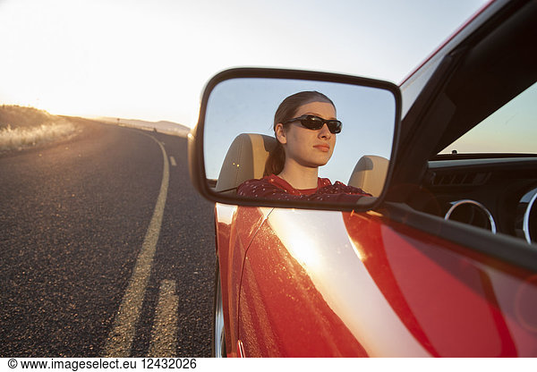 A young Caucasian woman reflected in side mirror of a convertible sports car on the highway.