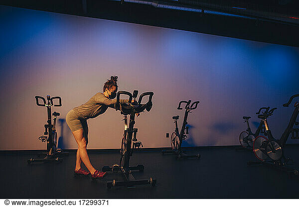 A young Caucasian woman stretches next to a stationary bike.