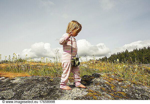 A young child in pajamas with a camera walks on a mossy rock