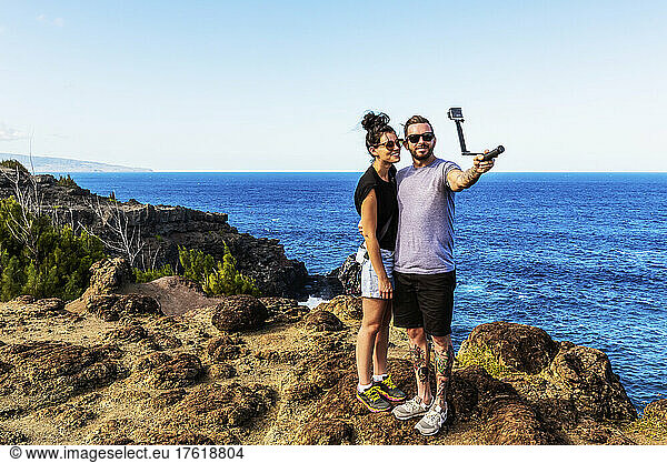 A young couple stands on the shore at Nakalele Point taking a self-portrait with the island's coastline and the Island of Molokai in the background; Maui  Hawaii  United States of America