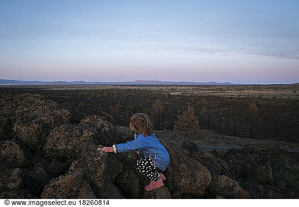 A young girl climbs rocks overlooking a vast desert at dusk