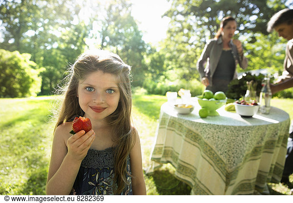 A Young Girl Holding A Large Fresh Organically Produced Strawberry Fruit. Two Adults Beside A Round Table.