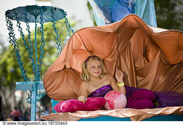 A young girl in a seashell at a parade in Santa Barbara. The parade features extravagant floats and costumes.
