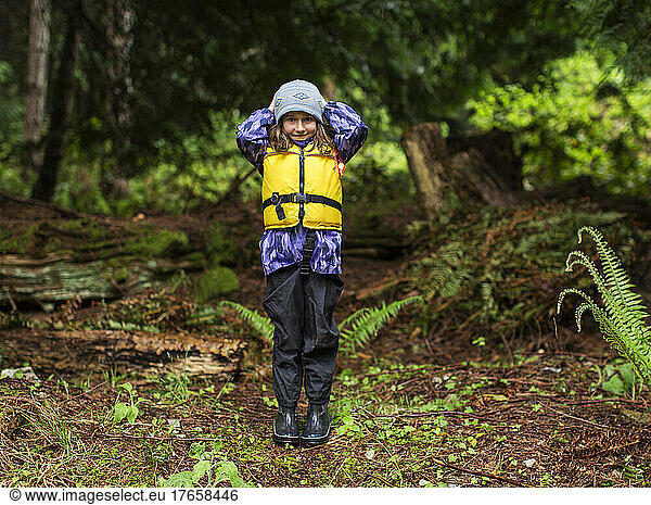 A young girl in rain gear and life jacket stands in the forest