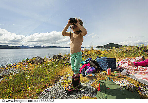 A young girl looks through binoculars on a mossy rock overlooking bay