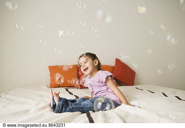 A young girl with brown eyes and dark hair in bunches sitting on a bed laughing. Bubbles floating in the air.