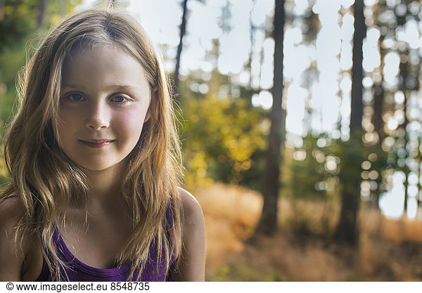 A young girl with long blonde hair in woodland in the fresh air  looking at the camera.
