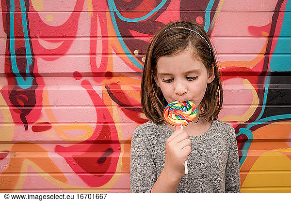 a young girls eats a huge rainbow lollipop in front of a colorful wall
