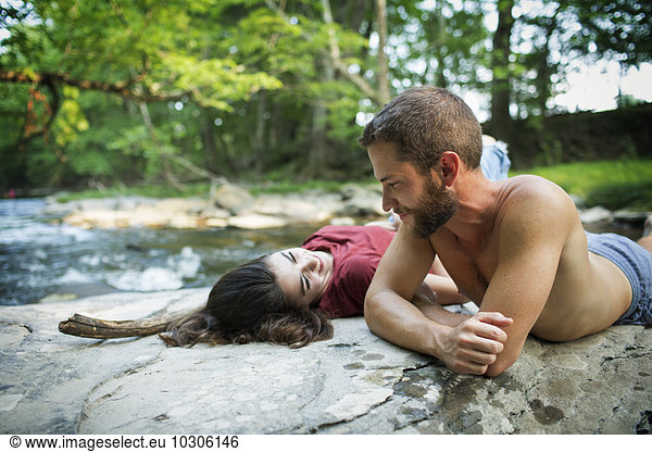 A young man and woman lying on the rocks on a river bank.