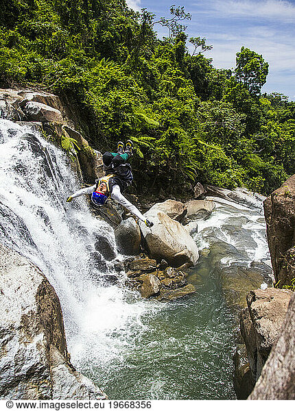 A young man does a back flip off a rocky outcrop into a swimming hole during a river adventure on a sunny day in Puerto Rico.