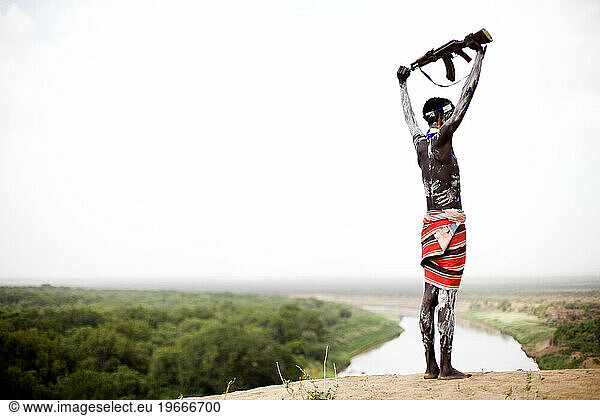 A young man holds his kalashnikov rifle over his head while overlooking the Omo river in the remote Omo Valley of Ethiopia.