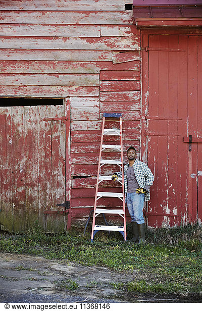 A young man in working clothes and cap carrying a ladder across a farm yard.