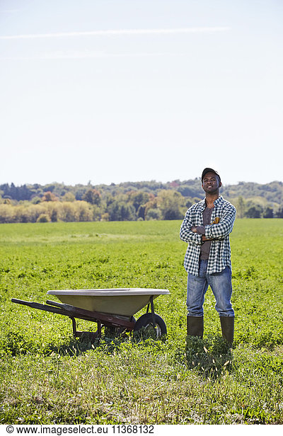 A young man in working clothes standing in a crop field with arms folded next to a wheelbarrow.
