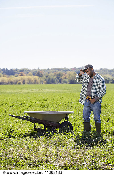 A young man in working clothes standing in a crop next to a wheelbarrow touching the brim of his cap.