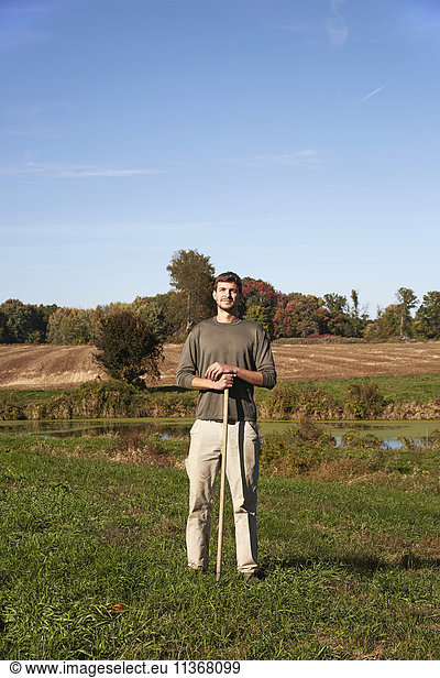 A young man in working clothes standing in a field  holding a tool.