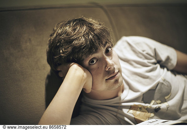 A young man lying on a sofa  resting his head on his elbow and looking at the camera.