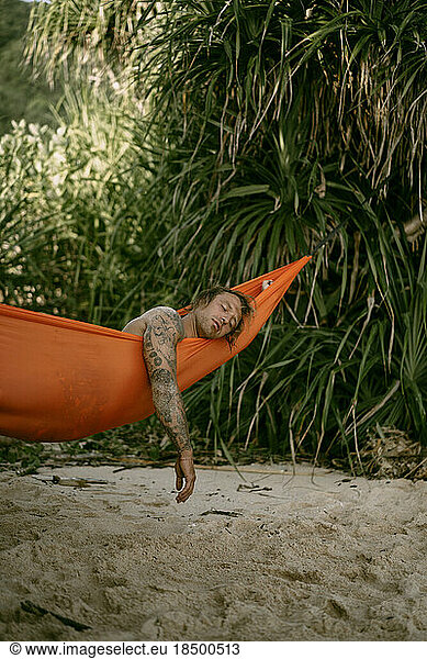 A young man rests and chills in an orange hammock on the beach n