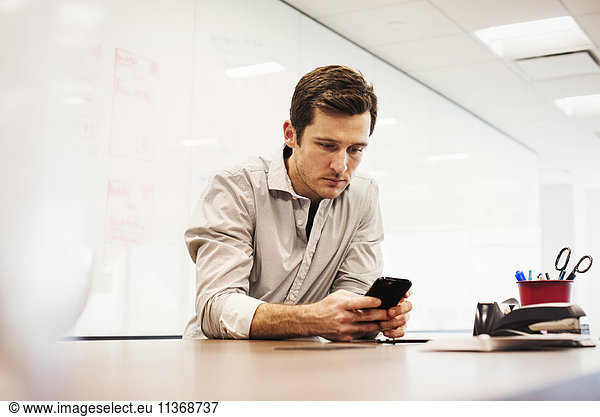 A young man sitting in a classroom at a table and looking down at a cellphone.