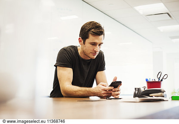 A young man sitting in a classroom leaning on a desk looking down at a cellphone.