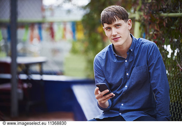 A young man sitting outdoors holding a cellphone and looking into shot.