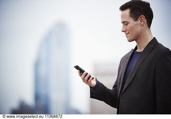A young man standing on a rooftop looking down at a cellphone.