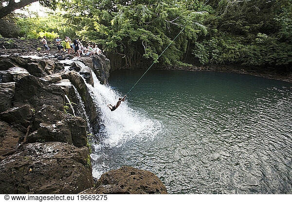A young man swings over a waterfall on a rope swing as a crowd enjoys watching the show.