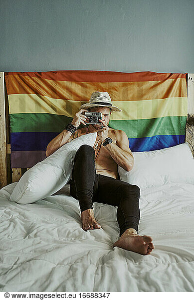 A young man with no shirt and hat in bed taking pictures with an LGBTQ rainbow flag