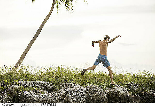 A young man without shirt jumps on rocks at near palm tree at water