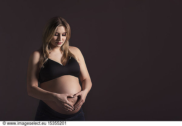 A young pregnant woman holding her belly in a studio and looking down while making a heart shape over her unborn child; Edmonton  Alberta  Canada