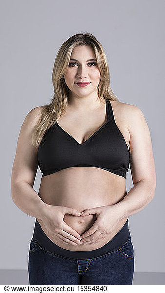 A young pregnant woman holding her belly in a studio and posing for the camera while making a heart shape over her unborn child; Edmonton  Alberta  Canada