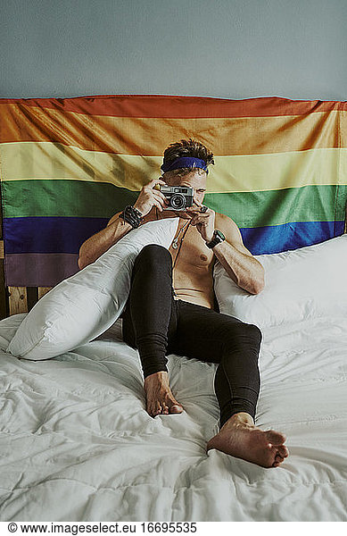 A young shirtless man in bed taking pictures with the LGBTQ rainbow flag