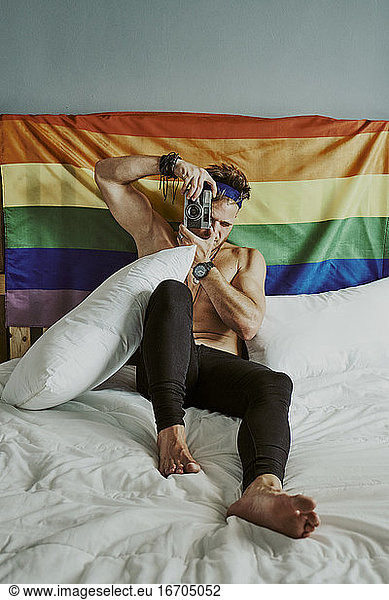A young shirtless man in bed taking pictures with the LGBTQ rainbow flag behind