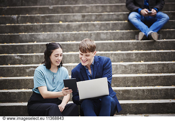 A young woman and a young man sitting on a flight of steps outdoors looking at a laptop together.