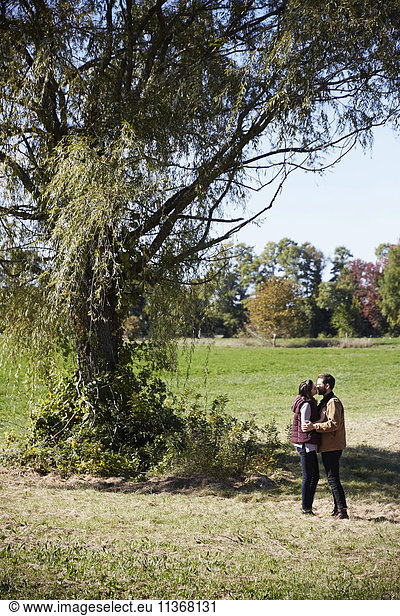 A young woman and man standing next to a tree in a field  kissing.