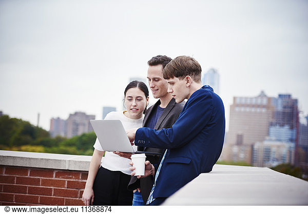 A young woman and two young men standing on a rooftop looking down at a laptop computer together.