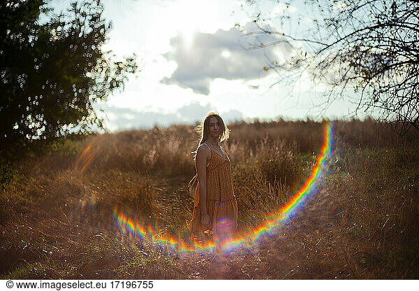 A young woman in a dress stands in a hay field with a lens flare highlights her frame