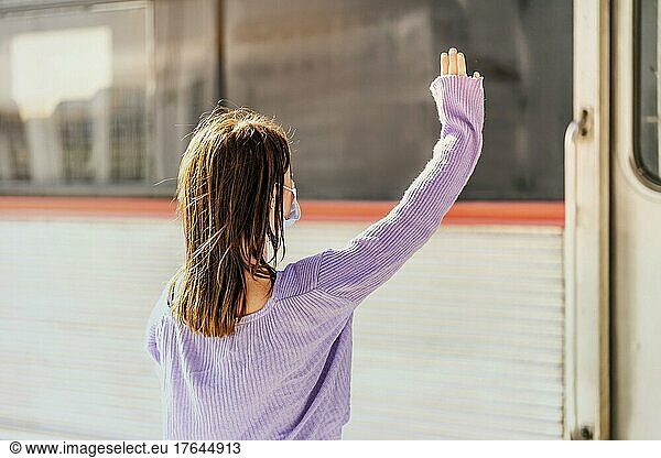 A young woman in a mask waves to somebody in the train from the platform of the railway station