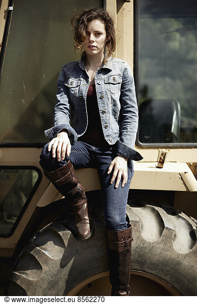 A young woman in denim jacket and boots on the hood of a tractor.