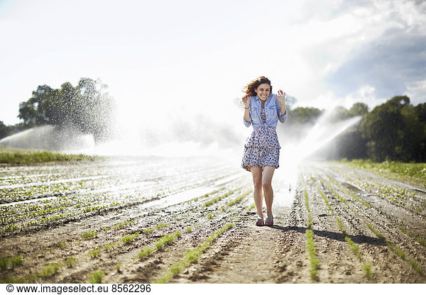 A young woman in denim jacket standing in a field  irrigation sprinklers working in the background.