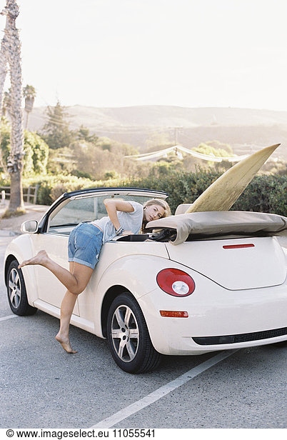 A young woman in denim shorts leaning into a convertible car with a surfboard in the back seat.