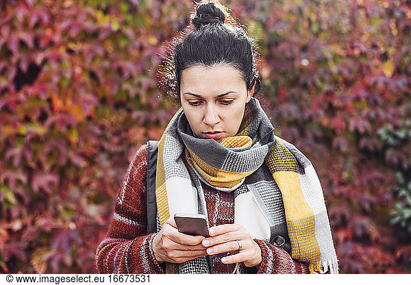 A young woman in scarf looking at her cell phone  red autumn leaves