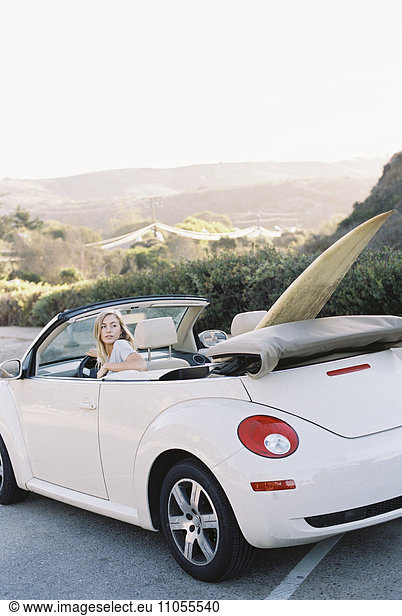 A young woman in the driving seat of a convertible with a surf board in the back seat.