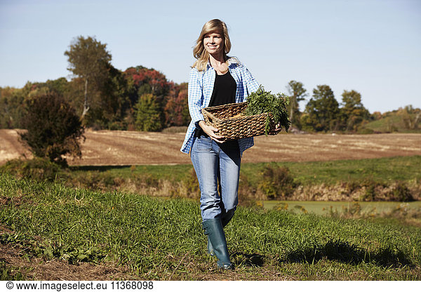 A young woman in working clothes walking across a field  holding a basket of crops.