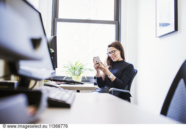 A young woman sitting at a desk in an office looking down at a cellphone.