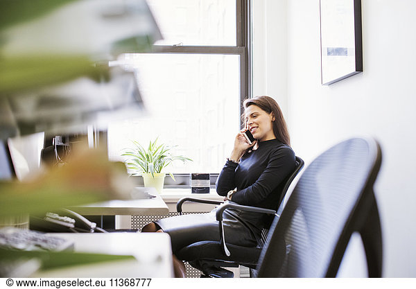 A young woman sitting at a desk with a cellphone to her ear.