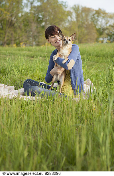 A Young Woman Sitting In A Field  On A Blanket  Holding A Small Chihuahua Dog.