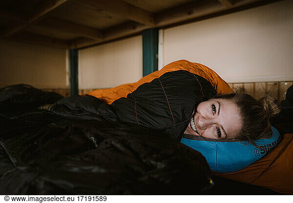 A young woman smiles laying inside her sleeping bag inside ski cabin