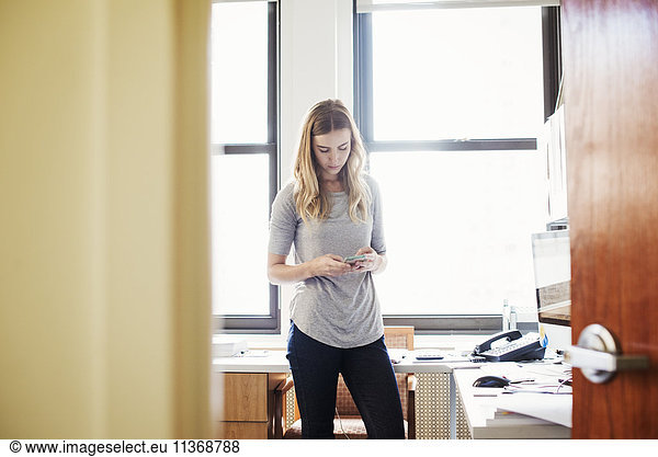 A young woman standing in an office looking down at her cellphone.