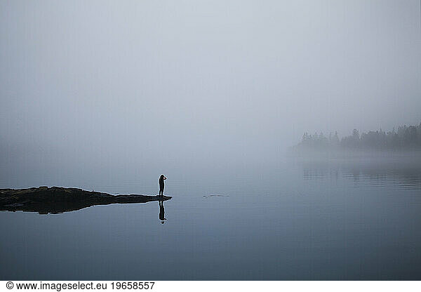 A young woman walks on the edge of a rock overlooking a foggy  calm body of water.