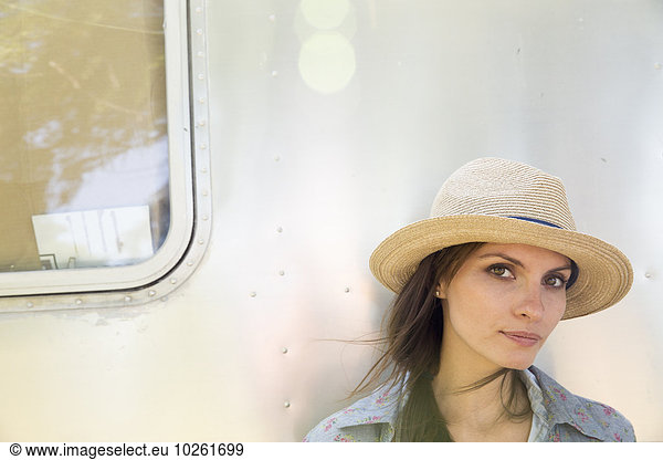 A young woman wearing a hat sitting in the shade of a silver coloured trailer.
